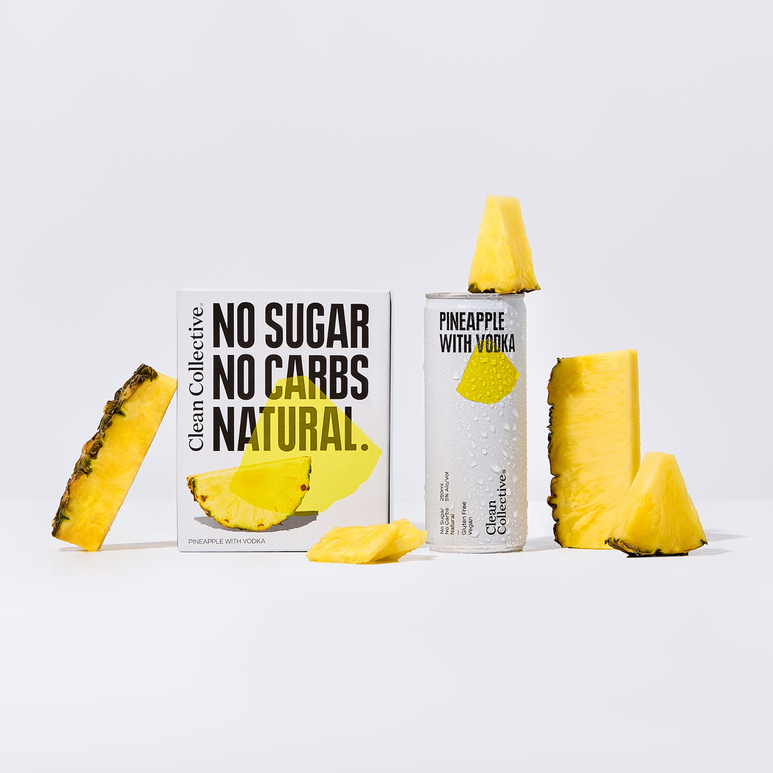Two cans of Clean Collective Pineapple with Vodka set against a light background with fresh pineapple chunks and slices. The front can highlights "NO SUGAR, NO CARBS, NATURAL".